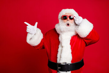 Photo of retired old man grey beard open mouth hold specs direct finger empty space show great solution wear santa x-mas costume gloves coat sunglass headwear isolated red color background
