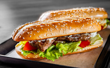 Two long submarine sandwiches with meat, cheese, bacon, tomatoes, lettuce, cucumbers and onions on black wooden table background