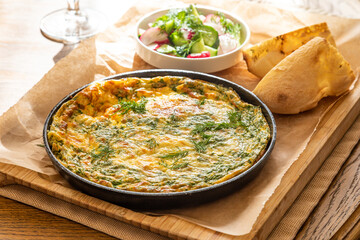 Omelet in a frying pan with parsley and dill. Serve with fresh bread and fresh vegetables.