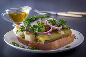 Sandwich with herring fillets, onions, pickled cucumber, egg and herbs on a white plate on a dark table. Traditional Scandinavian street food. Close-up
