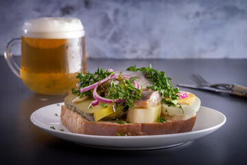 Sandwich with herring fillets, onions, pickled cucumbers, eggs and herbs on a white plate on a dark table against the background of a glass of beer with foam. Traditional Scandinavian street food