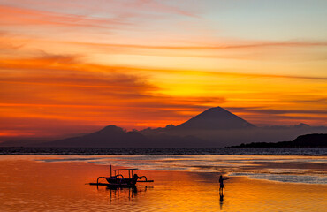 Silhouette of a person and a small traditional boat against sunset over the sea with a volcano in the distance