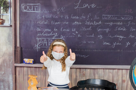 Life during coronavirus pandemic. girl in white blouse with medical protective mask showing thumbs up on chalkboard background. back to school concept. schoolgirl wearing facial mask. - Powered by Adobe