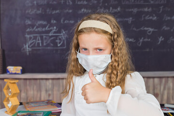 Life during coronavirus pandemic. girl in white blouse with medical protective mask showing thumbs up on chalkboard background. back to school concept. schoolgirl wearing facial mask.