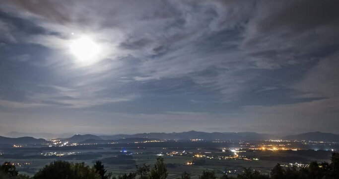 Elevated aerial view of night time lapse of clouds moving fast over flat basin and mountains in Slovenia. Moon over landscape. Villages illuminated by lights. Static wide shot
