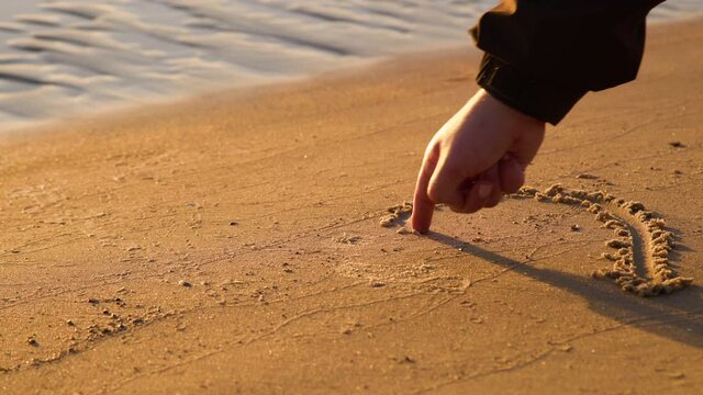 Close up view of woman hand drawing heart with finger on the sand at the beach. Romantic love symbol at tropical seashore at sunset