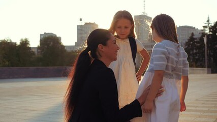Close up good looking brunette mum woman parent embracing two little kid sisters pupil on schoolyard. Mother talking smiling pretty daughters, sends off school girls with backpacks on sunset outdoor