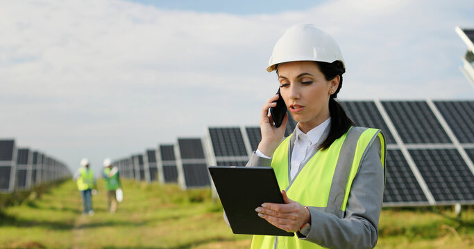 Portrait of female electrician engineer in safety helmet and uniform using tablet and talking on phone checking solar panels.