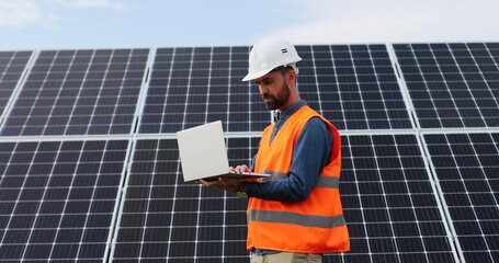 Portrait of electrician in safety helmet and uniform using laptop checking solar panels. Male...