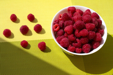 Raspberries in a white bowl on a green background. Ripe berries are scattered.