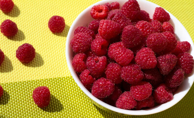 Raspberries in a white bowl on a green background. Ripe berries are scattered.