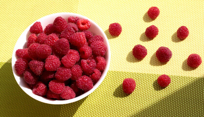Raspberries in a white bowl on a green background. Ripe berries are scattered.
