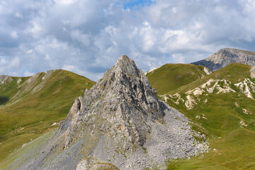 Fototapeta premium Rocky mountain peak, which rises in the middle of the prairie, in the Alps, between the grass on the slopes of the neighboring mountains and the cloudy sky with small blue glades. Gray rock peak.