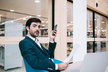 Portrait of Indian employee holding corporate report and cellphone technology and looking at camera during working process, formally dressed professional with mobile device and papers posing indoors