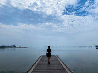 In the background of blue sky and white clouds, the back of a beautiful woman on the plank road on the lake surface