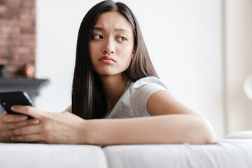 Image of unhappy asian woman using mobile phone while sitting on sofa