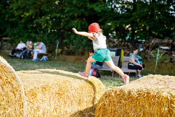 Little toddler girl having fun with running and jumping on hay stack or bale. Funny happy healthy child playing with straw. Active outdoors leisure with children on warm summer day. Kids and nature