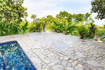An open and empty terrace of beige natural stone next to a blue pool surrounded by green plants...