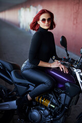 Fototapeta na wymiar A girl with red hairs posing with dark urban motorcycle