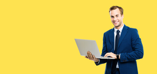 Portrait image - smiling businessman in blue confident suit, working with laptop, isolated against yellow color background. Handsome young man at studio concept picture. Copy space for text.