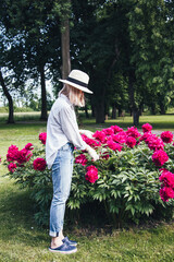Peony, garden, pink, summer, jeans, shirt, hat, woman. Background with peonies and woman in the garden. Pretty woman with blonde colored hair near a big peonies bush.