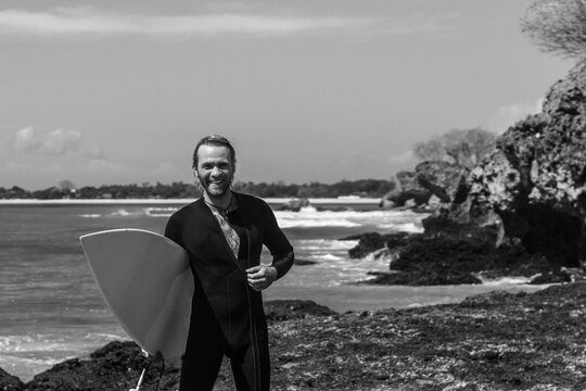 Black And White Portrait Of Handsome Man Surfer In Black Wetsuit, Holding White Surf Board  And Blue Ocean, Cliff, Rocks Behind..