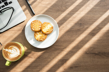 Top view of wood office desk table with cup of latte coffee, dish of almond cookies and office supplies with sun light. Life at work in the morning concept.