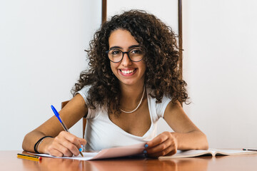 Young student girl with glasses smiling with curly brunette hair writing on a paper on a wooden table with a book, glasses and pens