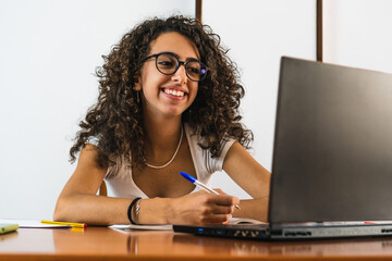 Young student girl with glasses smiling with curly brunette hair looking at a laptop on a wooden table with a book, glasses and pens