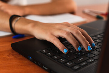 Detail of girl's hand typing on a black laptop on a wooden table. Her nails are painted blue.