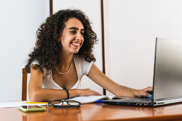 Young smiling student girl with curly brunette hair using a laptop on a wooden table with a book, glasses and pens