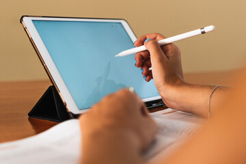 Detail of student girl hand picking up a digital pen using a tablet at a table with books and pens