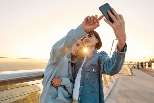 Lesbian couple standing on the bridge, posing while taking a selfie picture, watching the sunrise together