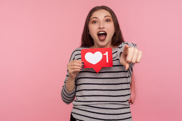 Hey you, click Like and follow my blog! Portrait of blogger woman in striped sweatshirt holding social media Heart button and pointing finger to camera. indoor studio shot isolated on pink background