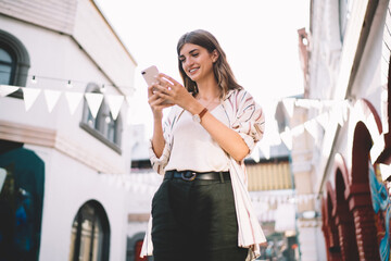 Smiling female messaging at city urbanity and using smartphone device with 4g wireless connection, happy woman dressed in casual clothes using cellphone technology for online chatting with followers