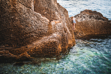 A girl climbs the stones near the coastal sea cliff - clear clear water