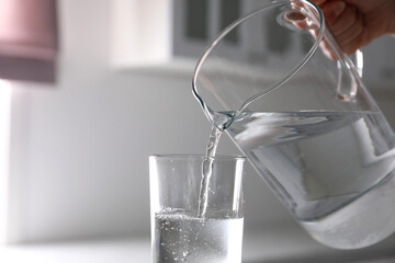 Woman pouring water from jug into glass in kitchen, closeup