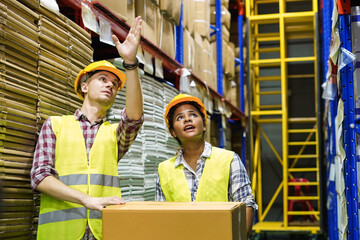 Warehouse manager with safety helmet pointing towards shelf in warehouse and talk to worker to assign job