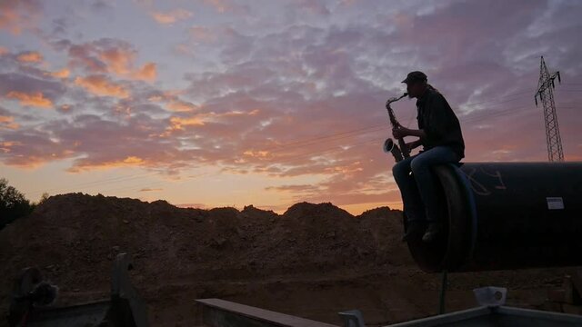 Mann sitzt auf Pipeliner&ouml;hre mit Saxophon vor Sonnenuntergang mit Feldern im Hintergrund