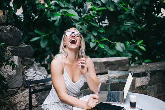 Excited female freelancer in classic glasses holding cellular technology and laughing near table in street cafe, toothy Caucasian woman 30s grinning during distance working on business project