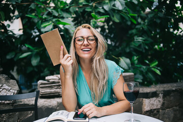Happy successful female reader with interesting literature book taking rest in street cafe during...
