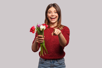 Flowers for you! Portrait of cheerful happy brunette girl smiling, holding tulips bouquet and pointing at camera. Congratulations on spring holidays. indoor studio shot isolated on gray background