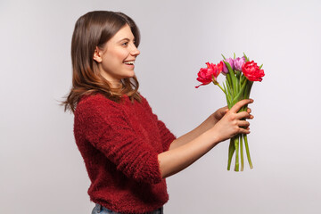 Side view portrait of cheerful happy brunette girl giving flowers bouquet and smiling excitedly. Congratulations on spring holidays, 8 March Women's Day. indoor studio shot isolated on gray background