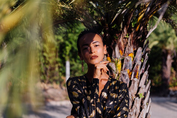 Fashion exotic portrait of slim tanned fit short dark hair woman with bronze make up in black golden long cape by palm tree, shadows falling on face and body from palm leaf. 