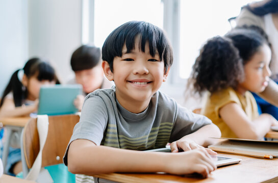 Cute little Asian boy smiling looking at camera and using laptop in computer class at the elementary school. Education, school, technology and internet concept.