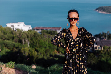 Fototapeta premium Portrait of beautiful luxury look fit tanned slim woman in black golden cape and sunglasses on top of mountain with amazing sea view in Thailand. 