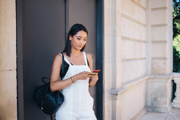 Pensive caucasian millennial woman reading income message on smartphone standing outdoors on street, young 20s hipster girl watching video from blog on mobile phone connected to 4G internet