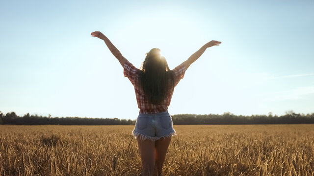Happy woman walking on a wheat field with open arms at the bright sun.