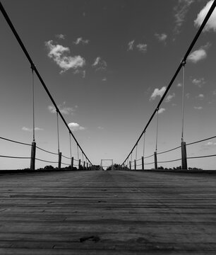 Low angle black white suspension bridge with clouds in sky wooden plank walkway traversing colorado river in Texas USA.