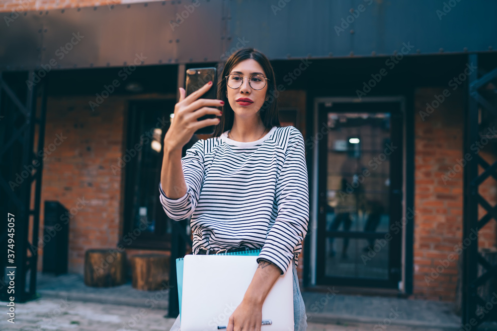Poster latino student with netbook posing at urban setting for making web selfie content via front smartpho